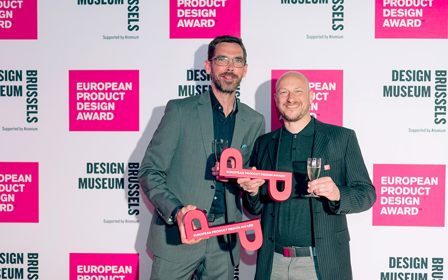 Due vincitori tengono in mano i loro trofei dell’European Product Design Award durante la cerimonia di premiazione, davanti a un pannello con il logo del premio